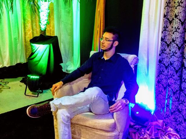 Un hombre con una apariencia más atractiva, con gafas, camisa oscura, pantalones blancos, sentado en una silla, con los brazos relajados, luces verdes y azules, cortinas negras y blancas, papel tapiz con motivos florales y un foco verde..