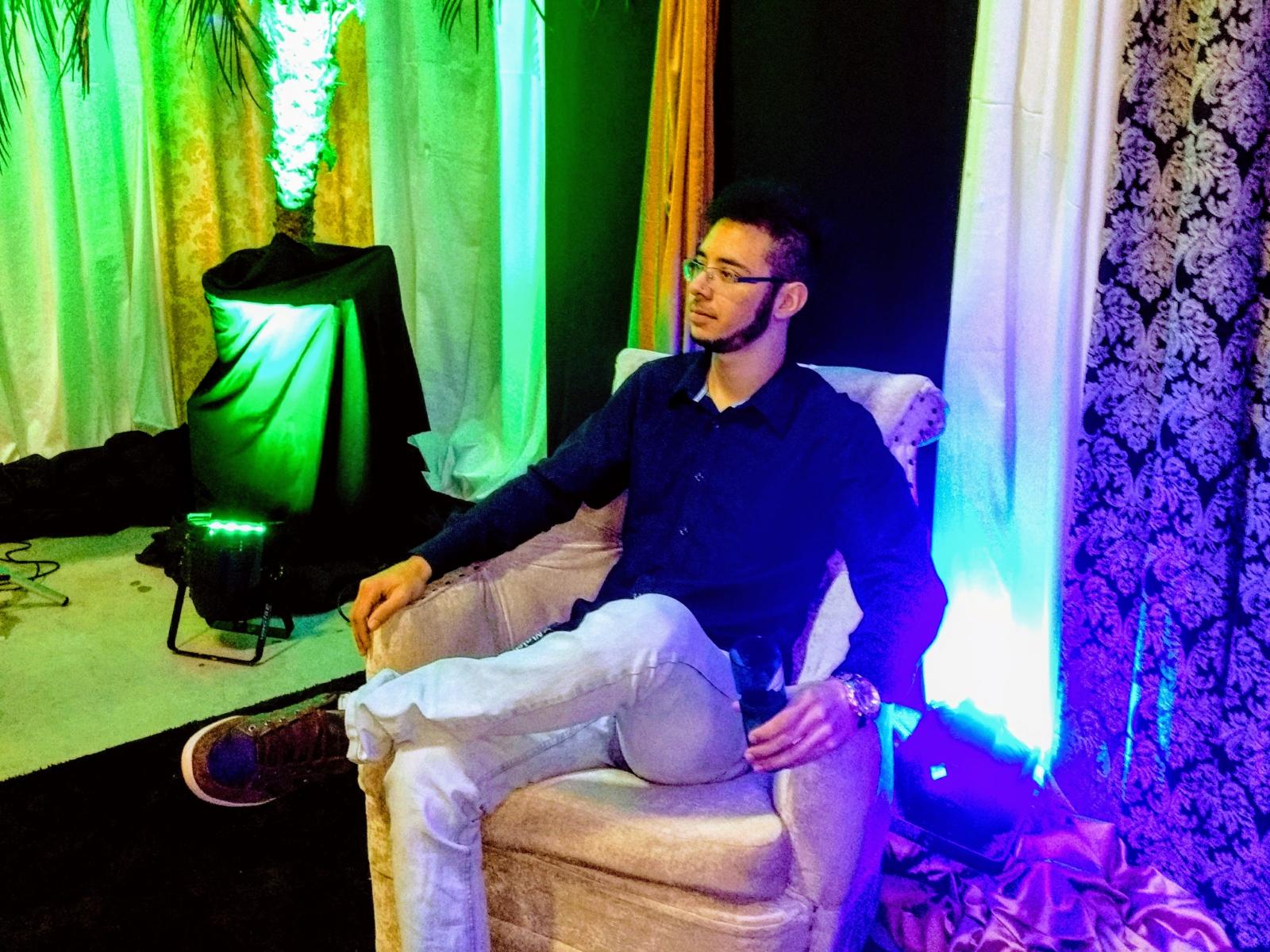 Un hombre con una apariencia más atractiva, con gafas, camisa oscura, pantalones blancos, sentado en una silla, con los brazos relajados, luces verdes y azules, cortinas negras y blancas, papel tapiz con motivos florales y un foco verde..