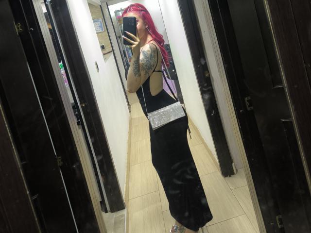 SammyViolet with pink hair, black backless dress, silver belt, tattooed arm, holding phone, mirror selfie, showing ass, in a dark hallway with wooden floors.