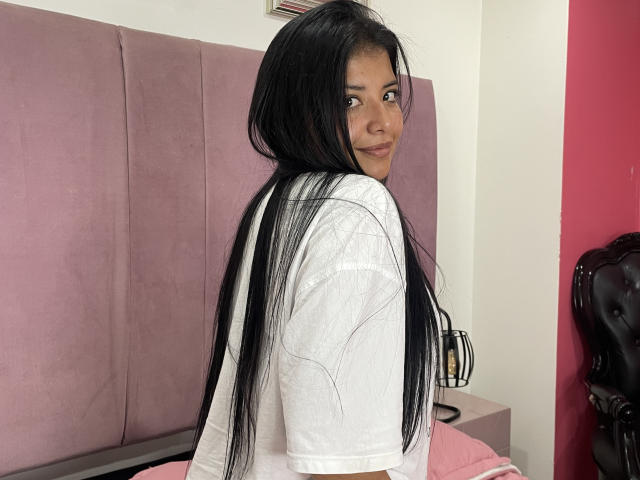 JuLietaBlasCo, com cabelos pretos longos, camisa branca, cama rosa, olhando para trás com um sorriso discreto, mostrando levemente o bumbum. Seus olhos escuros estão fixos na câmera..