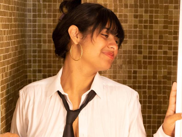 AnanyaSyed, con el cabello oscuro recogido en un moño, vestida con una camisa blanca holgada y una corbata negra, sonriendo ligeramente, tocando una pared con un patrón de azulejos dorados, y llevando grandes aros de oro..