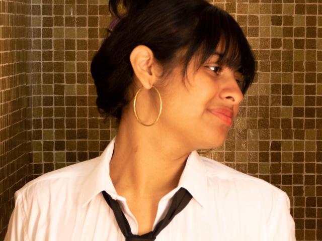 AnanyaSyed, con cabello oscuro, aros grandes, una camisa blanca, una corbata negra y una ligera sonrisa, mirando hacia un lado. Tiene un pequeño moretón en el cuello..