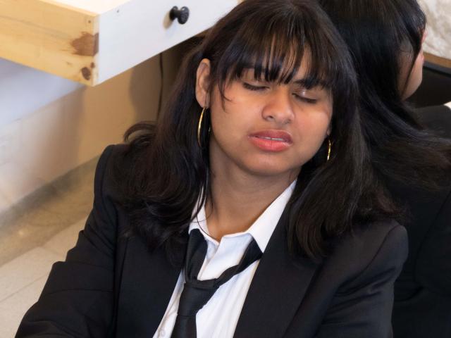AnanyaSyed, com os olhos fechados, cabelo preto e franja, vestindo um terno preto, camisa branca e gravata preta, usa pequenos brincos de ouro e tem uma expressão ligeiramente emburrada e séria..