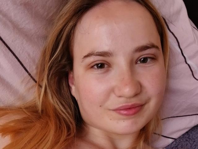 SofieBloom has light skin, brown eyes, and blonde hair with orange tips, smiling slightly with a small mole on her right cheek, lying on a pink pillow.