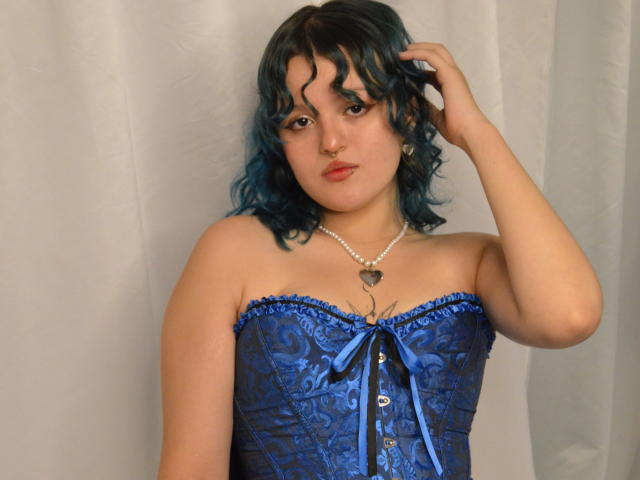 StephyStar with blue curly hair, wearing a blue lace corset with a ribbon, pearl necklace, and heart pendant, touching her hair, looking at the camera.