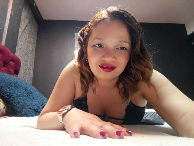 BiancaMorettii with brown wavy hair, red lipstick, black lace bra, pink nail polish, wristwatch, lying on bed, smiling, hands on bed, small tattoo on chest.