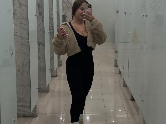 NatSorian with medium tits, black lace top, and tight black pants, wearing a beige fuzzy jacket, taking a mirror selfie in a marble bathroom, holding a pink phone.