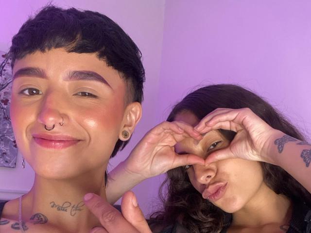 Riley e Alexa, con capelli neri corti, piercing al naso e tatuaggi, sorridono a sinistra. Alex, con capelli castani ondulati, forma un cuore con le mani e stringe le labbra a destra..