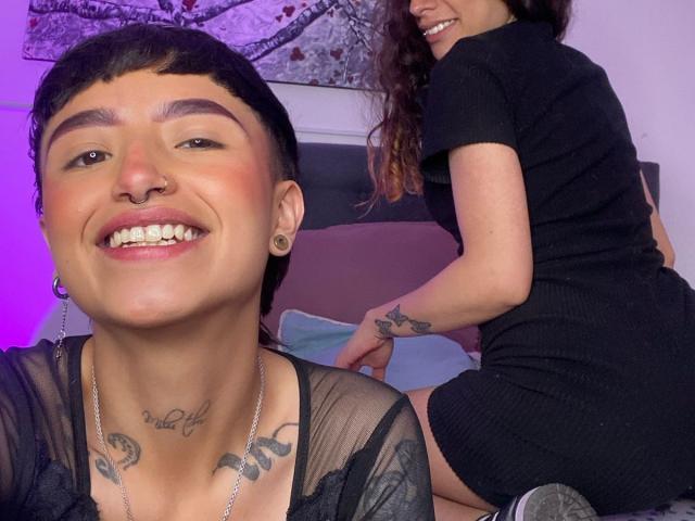 RileyAndAlexa con capelli neri corti, piercing al naso e espansori alle orecchie, sorridenti, che mostrano tatuaggi, indossano una maglia nera trasparente, una collana d'argento e un vestito nero, sedute sul letto con RileyAndAlexa che sorride..
