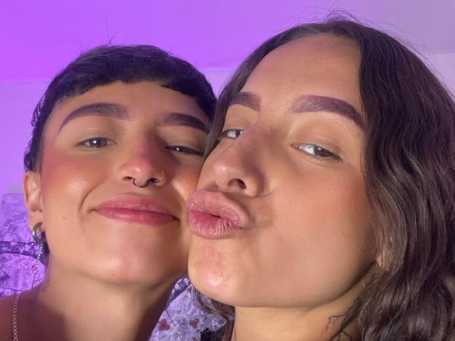 RileyAndAlexa, both with cute faces, short brown hair, and nose rings, are kissing their pouty lips together. Purple lighting highlights their skin. RileyAndAlexa are making kissy faces.