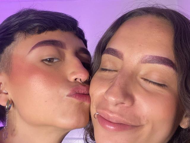 Die Gesichter von Riley und Alexa, die sich küssen, Riley hat einen Nasenring, Ohrringe und dunkle Augenbrauen, Alexa hat einen Lippenpiercing, beide haben rosig-rote Wangen, braune Augen und geschlossene Lächeln..