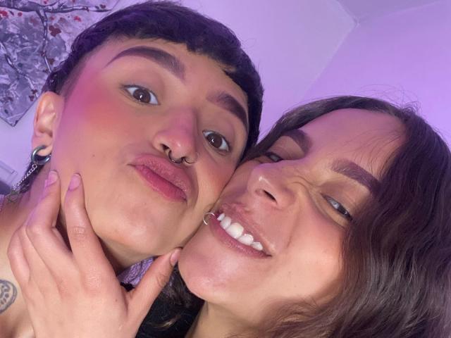 RileyAndAlexa, both with short hair, have a nose ring and lip ring respectively. Riley's hand is on Alexa's cheek, smiling with visible teeth. Pinkish purple light.