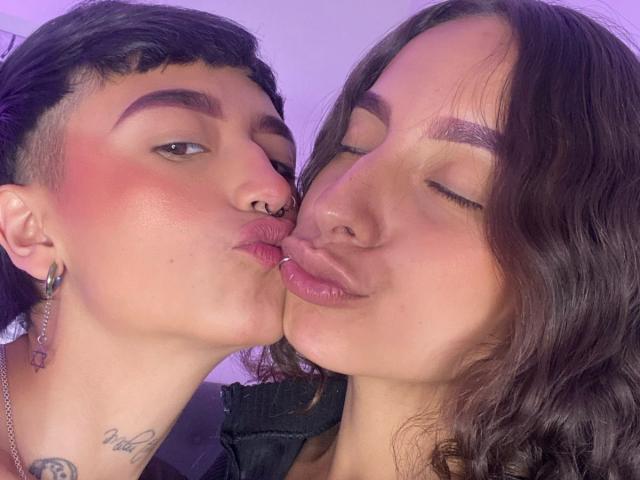 RileyAndAlexa are kissing with puckered lips, both with pink blush, nose rings, and tattoos. Riley has short black hair, Alexa has wavy brown hair. Both have shiny, glossy lips.