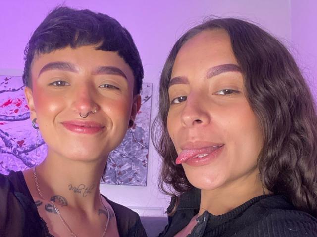 RileyAndAlexa, two chicks with short black hair and tattoos, smiling with nose rings and earrings, one with tongue out, in black shirts, under purple lighting.