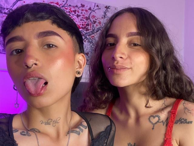 Riley und Alexa, beide mit kurzen, schwarzen Haaren und Nasenring, strecken die Zunge heraus. Alexa hat welliges, braunes Haar, ein rotes Spitzenoberteil, Tattoos und einen Nasenring und lächelt. Beide haben sichtbare Tattoos..