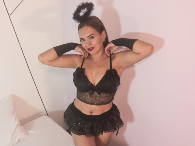 Rachel, uma escocesa de cabelos escuros, vestindo um sutiã de renda preta, uma saia curta e braceletes, usa uma tiara preta e fofa, está em pé com os braços levantados, mostrando seus seios e nádegas..