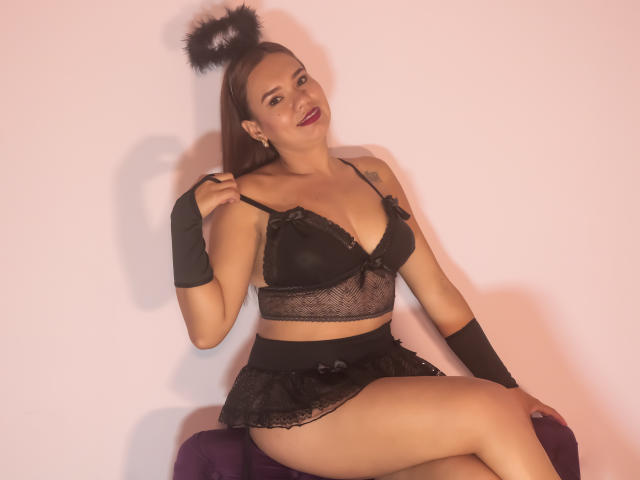 Rachel, una chica con lencería negra, un sujetador de encaje negro y tanga, muñequeras negras, orejas de gato negras y una sonrisa encantadora, sentada sobre un paño morado, mostrando su trasero y sus pechos..