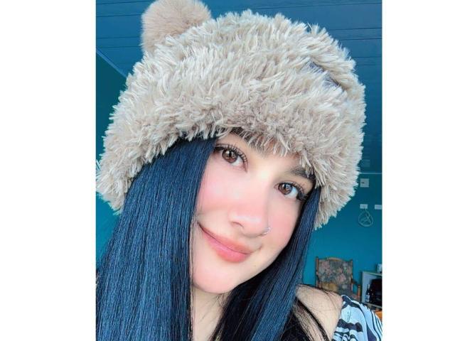 AdharaBritney with long blue hair, smiling, wearing a fluffy beige bear hat, nose ring, and a black-and-white striped shirt. Her brown eyes are cute.