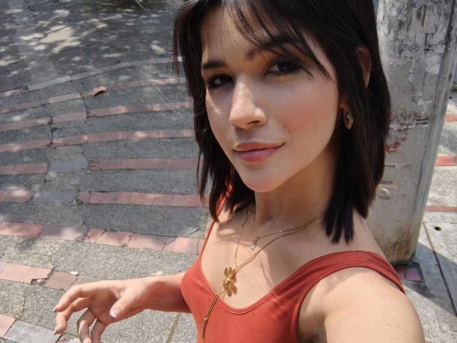ScarlletZhen with short black hair, brown eyes, and pink lips, wearing a red tank top and gold necklace with a cross, standing by a tree, looking at the camera.