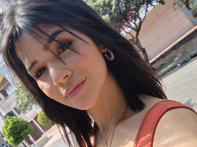 ScarlletZhen tem cabelo escuro, olhos castanhos e lábios rosados, veste uma blusa vermelha, exibindo um sorriso adorável com pequenos brincos. A luz do sol realça seu rosto..