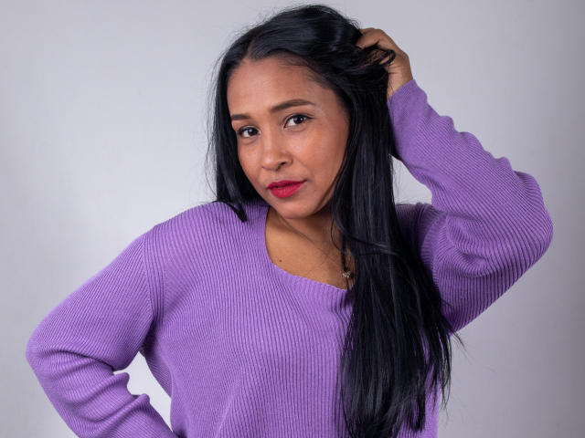 VictoriaSandoval with long black hair, wearing a purple rib-knit sweater, hand in hair, red lipstick, and a slight smile, against a plain gray background.