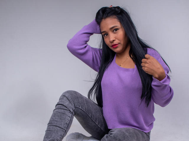 VictoriaSandoval with long black hair, purple sweater, and gray jeans, sitting, hand in hair, looking at camera, slight smile, red lipstick, necklace, casual pose, confident.