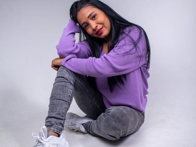VictoriaSandoval with long black hair, purple sweater, gray jeans, and white sneakers, sitting with knees up, hand in hair, smiling, red lipstick, and brown eyes.