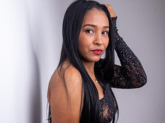 VictoriaSandoval with long black hair, wearing a black, sheer, long-sleeve top with small white dots, hand in hair, looking at camera, red lipstick, cute, sexy.