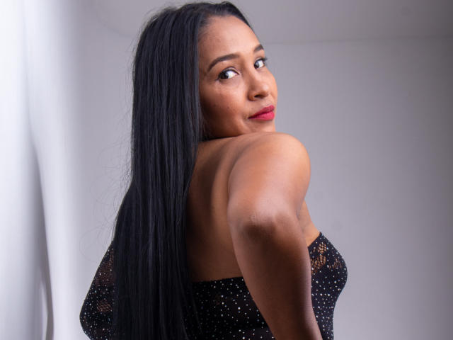 VictoriaSandoval with long black hair, red lipstick, and dark skin, looking over her shoulder at the camera, wearing a black, sheer, polka-dotted top, showing her bare, small ass.