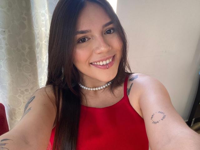 SophiaMarx with long brown hair, smiling, red dress, pearl necklace, tongue piercing, tattoos on arms, fair skin, cute, sexy, showing tits, holding phone for selfie.