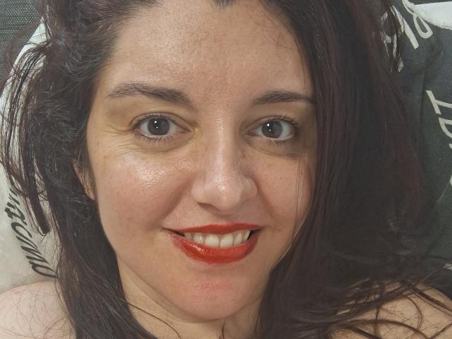 La Française Jessy, avec du rouge à lèvres, souriante, montrant ses dents, avec des yeux marron et des cheveux châtain foncé légèrement en désordre, est allongée et regarde l'appareil photo. Son visage a quelques taches de rousseur..