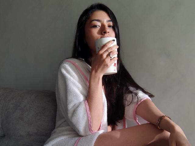 CanelleGarces with long black hair, sipping from a white mug, wearing a pink-trimmed white robe, sitting on a gray sofa, wearing a gold bracelet, looking relaxed.