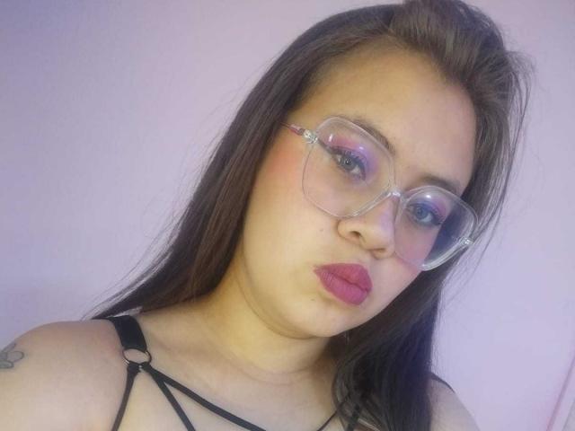 Isabella Reid, com cabelos castanhos longos, batom rosa, óculos de grau e uma blusa preta com tiras, aparece na foto mostrando seus seios. Ela tem uma tatuagem no ombro esquerdo..