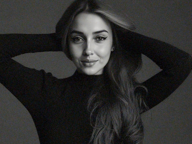 IsaBlonde69 with hands in her long, wavy hair, wearing a black turtleneck, has a subtle smile and is looking at the camera in a grayscale portrait.