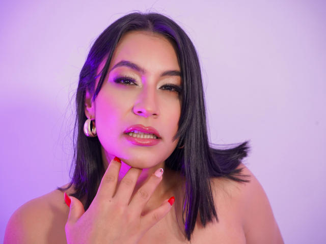 EmaFoxy has black hair, pink lips, red nails, and is touching her chin with her hand. She's wearing hoop earrings, and has a sexy, confident expression.