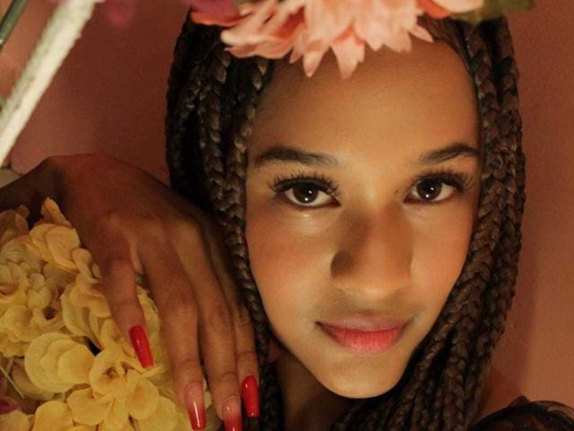 Ally Mills met donkere vlechten, een roze bloemenhoofddeksel, rode nagels, die gele bloemen vasthoudt, lachend, met bruine ogen en een schattig gezicht, in een warme, zacht verlichte selfie..