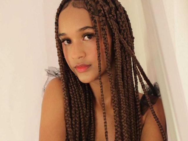 AllyMills with long brown braids, full pink lips, and dark eyes, wearing a black top, looking to the side with a subtle smile. Her ass is not visible.