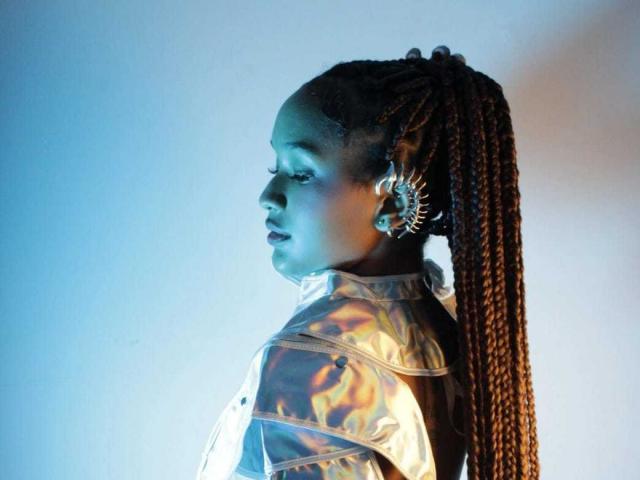 AllyMills with long brown braids, wearing a shiny metallic jacket, and a sunburst earring, looking to the side with blue lighting on her face.