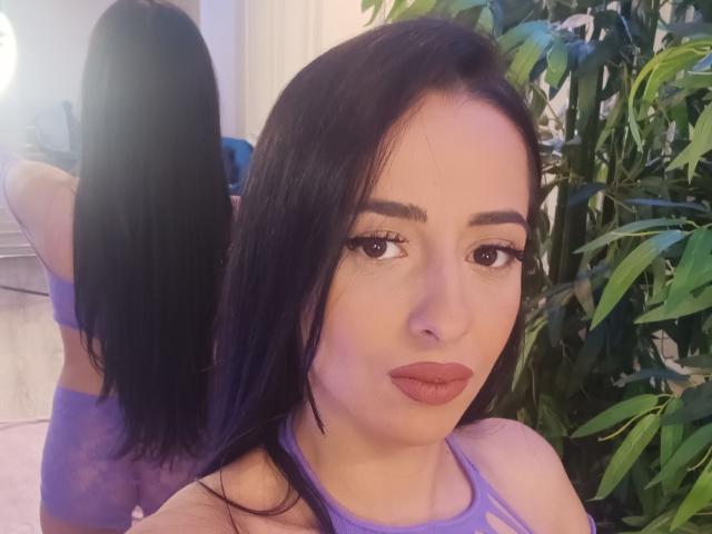 SallyMadison with long black hair, purple lace top, and pouty lips, standing in front of a mirror showing her back, reflecting her tits and ass. Potted plant on right.