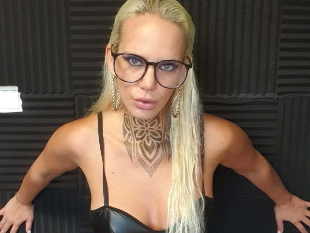 NiciXFetisch-hot with blonde hair, black glasses, and a large chest tattoo, wearing a black leather top, leaning forward with arms on a black textured wall.