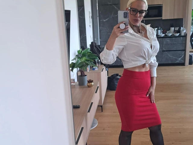 NiciXFetisch-hot with blonde hair, glasses, white button-up, tied at the midriff, red skirt, black stockings, showing her tits, holding phone, in a modern kitchen.