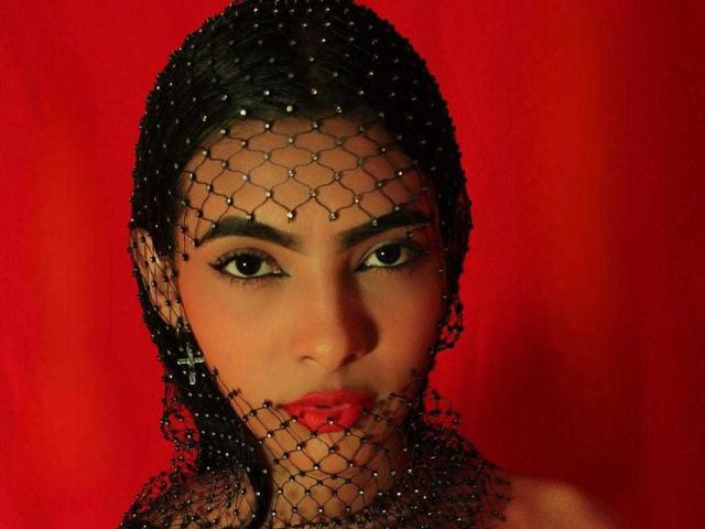 JoyFirestone has dark eyes, thick eyebrows, and red lipstick, wearing a black netted headpiece with tiny beads, looking serious against a red background.