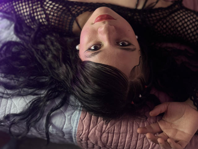 WhitneyLovee mit schwarzen Netzstrümpfen, liegt auf einem Bett, kopfüber, und schaut in die Kamera. Sie trägt rosafarbenen Lippenstift, schwarzes Haar und eine pinke, gesteppte Oberteil. Ihre Hände sind in der Nähe ihres Gesichts..