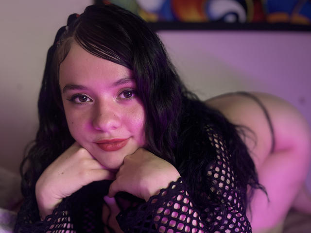 WhitneyLovee con capelli neri ondulati, un leggero riflesso rosa-viola sul viso, indossa una maglia a rete, le mani sul mento, guarda la fotocamera, con il sedere in vista attraverso uno slip, con un leggero sorriso, sdraiata sul letto..