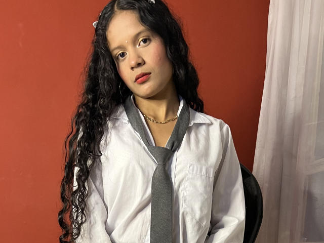 SharonLiam, con cabello negro largo y rizado, vestida con una camisa blanca y una corbata gris, lleva una pequeña cadena de oro y tiene pintados los labios de rojo, está sentada en una silla negra frente a una pared roja..