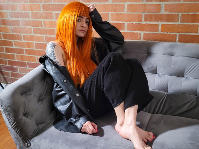 Amelie Sato, con cabello naranja, chaqueta negra estilo hombro descubierto, pantalones negros, tatuaje en el hombro izquierdo, sentada en un sofá gris, descalza, con la mano en el cabello, mirando a la cámara, suelo de madera..