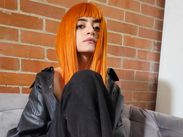 Amelie Sato, con un corte de pelo bob de color naranja brillante, mirando hacia abajo, vestida con pantalones negros y una chaqueta de cuero negra, sentada en un sofá gris, con una pared de ladrillos en el fondo..