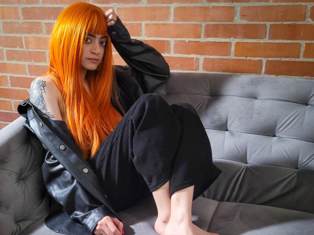 Amelie Sato mit leuchtend orangefarbenen Haaren, schwarzen Hosen und einem offenen schwarzen Hemd, sitzt auf einem grauen Sofa und zeigt ein Tattoo an ihrer rechten Schulter, barfuß..