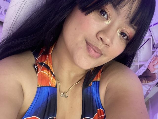 JulietaDuval with black hair, wearing glittery eyeshadow, a colorful halter top, and a gold necklace, smiling at the camera, showing her tits slightly.