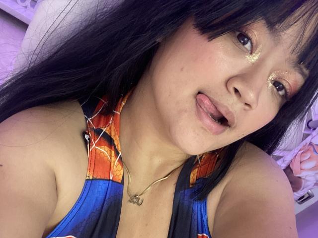 JulietaDuval with black hair, gold glitter eyeshadow, tongue out, wearing a blue and red Spider-Man tank top, necklace, and shiny skin, looking at the camera.